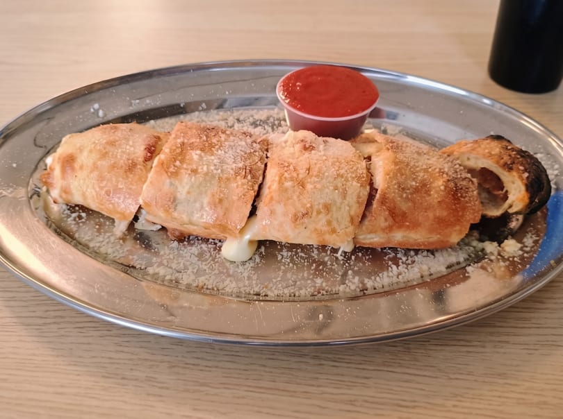 A sliced stromboli with melted cheese and parmesan on a metal plate with a side of marinara sauce.