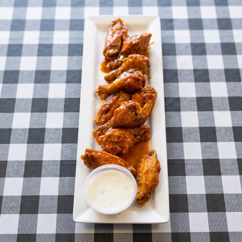 A long white platter of saucy chicken wings and a side of ranch dressing on a checkered tablecloth.