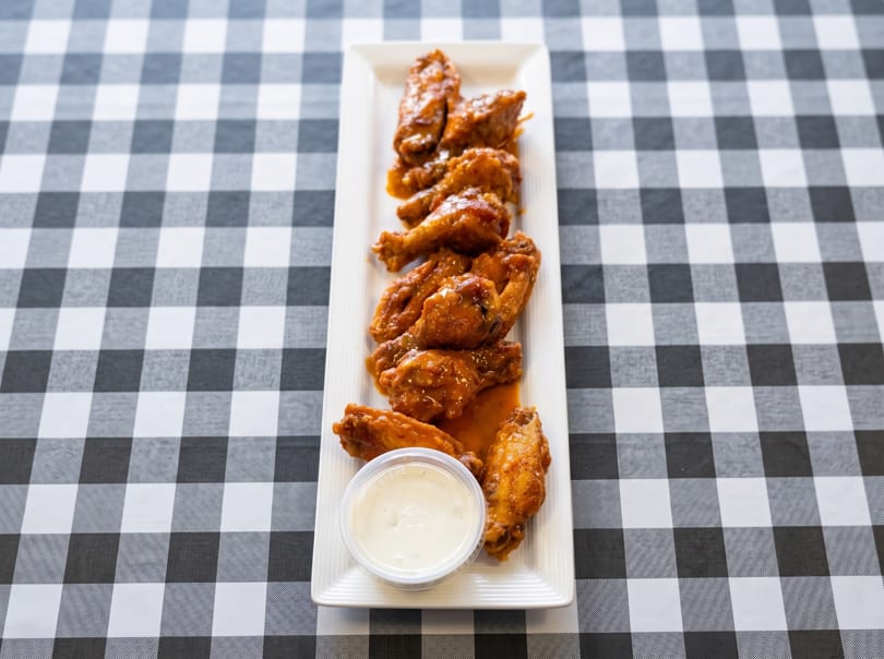 A long white platter of saucy chicken wings and a side of ranch dressing on a checkered tablecloth.