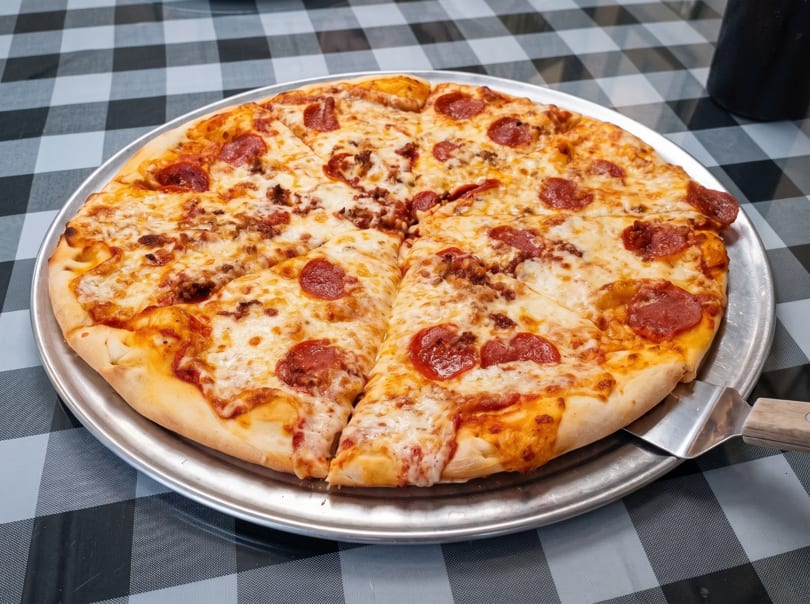 A whole pepperoni and sausage pizza on a metal tray, with one slice being served.